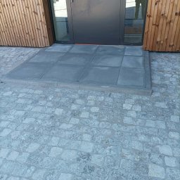 Katarzyna Zmiendak - Nowoczesne wejście do budynku z drewnianą elewacją, gdzie gładkie, ciemnoszare płyty betonowe tworzą próg, otoczony jasnoszarą, drobną kostką granitową.