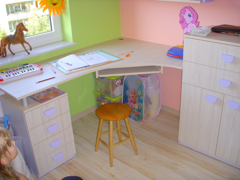 Pokój dziecięcy z biurkiem narożnym, krzesłem i szafką z szufladami, zabawki i dekoracje na ścianach.