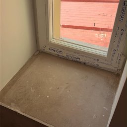 AngelHome - Zabrudzony parapet pod nowym oknem PVC z widoczną folią ochronną GEALAN, pokryty pyłem i resztkami budowlanymi, z czerwonym dachem w tle.