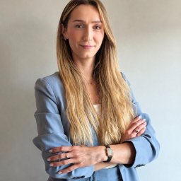 Usługi Oliwia Urban - Kobieta z długimi blond włosami w błękitnym garniturze, skrzyżowane ręce, delikatny uśmiech, zegarek na lewym nadgarstku, jasnoszare tło.