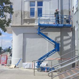 Bohdan Kozak - Pracownik na niebieskim podnośniku nożycowym wykonuje prace na elewacji jasnego budynku z ozdobnymi detalami. Widoczne są fragmenty fasady w trakcie renowacji, obok schody i chodnik.