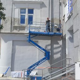 Bohdan Kozak - Pracownik na niebieskim podnośniku nożycowym wykonuje prace wykończeniowe na jasnej elewacji budynku, obok muralu z historyczną sceną uliczną.