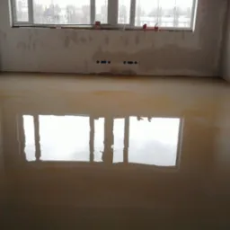 Wnętrze pokoju w trakcie remontu, z wylewką samopoziomującą na podłodze, ściany przygotowane do dalszych prac wykończeniowych, widoczne puszki elektryczne i okno z odbiciem na mokrej powierzchni.