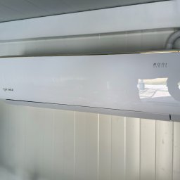 RUFI-KLIMA - Nowoczesny, biały klimatyzator ścienny Rotenso Roni Series ze złotym paskiem, zamontowany na białej, panelowej ścianie. Widoczna elastyczna rura instalacyjna.