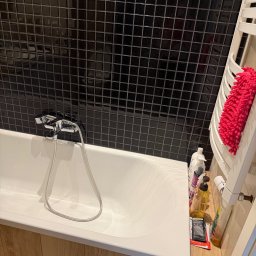 pani czystości - Czysta biała wanna z chromowaną baterią i ścianą z czarnych, błyszczących płytek mozaikowych. Obok wanny biały grzejnik drabinkowy z jaskraworóżową ściereczką.