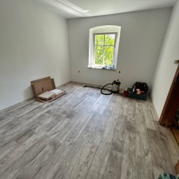 Firma Usługowa Kamil Lubiński - Jasny pokój z nowo ułożoną podłogą laminowaną w odcieniach szarości, białymi ścianami i łukowym oknem. Widoczne narzędzia i materiały do wykończenia.