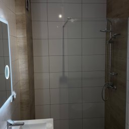 Przemek Rymer - Nowoczesna łazienka z prysznicem walk-in, białymi płytkami ściennymi i drewnopodobnymi panelami. Widoczna umywalka z chromowaną baterią i podświetlanym lustrem.