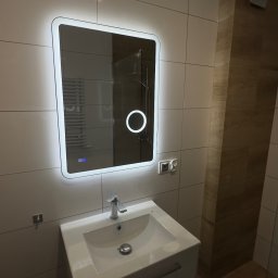Przemek Rymer - Nowoczesna łazienka z białą szafką pod umywalką i podświetlanym lustrem z dotykowym panelem. Jasne płytki, drewnopodobny element i widoczny grzejnik w odbiciu.