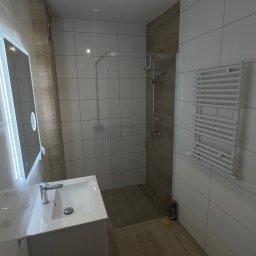 Przemek Rymer - Nowoczesna łazienka z białymi płytkami i drewnopodobnymi akcentami. Wnętrze obejmuje kabinę walk-in z deszczownicą, podwieszaną toaletę, szafkę z umywalką, lustro LED oraz biały grzejnik drabinkowy.