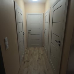 Przemek Rymer - Nowoczesny korytarz z jasnymi drzwiami wewnętrznymi z poziomymi frezami, drewnopodobną podłogą i okrągłą lampą sufitową. Beżowe ściany i klapa rewizyjna na suficie.