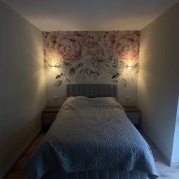 Przemek Rymer - Przytulna sypialnia z dużym łóżkiem, szarą pikowaną narzutą i tapicerowanym zagłówkiem. Ściana akcentowa z fototapetą w różowe piwonie, oświetlona dwoma kinkietami. Kryształowa lampa sufitowa...