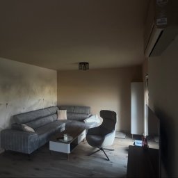 Przemek Rymer - Nowoczesny salon z szarą narożną sofą, designerskim fotelem i stolikiem kawowym. Ściana z delikatnym wzorem dmuchawców, na prawej ścianie klimatyzator. Jasna drewniana podłoga.