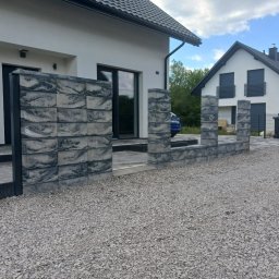 Bartek Domagała - Nowoczesne ogrodzenie z szarych, fakturowanych bloczków betonowych z czarnymi słupkami, prowadzące do wejścia białego domu z ciemnym dachem, z podjazdem z jasnego kruszywa.