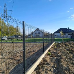 Bartek Domagała - Ciemnozielone ogrodzenie panelowe z betonową podmurówką, biegnące wzdłuż świeżo przygotowanego gruntu, na tle nowo budowanych domów i słupa wysokiego napięcia, pod czystym, błękitnym niebem.