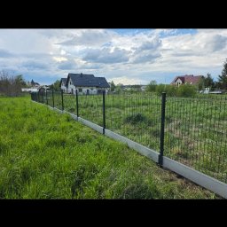 Bartek Domagała - Czarne ogrodzenie panelowe z szarą podmurówką betonową, biegnące wzdłuż zielonej, niezagospodarowanej działki. W tle nowoczesne domy pod częściowo zachmurzonym niebem.