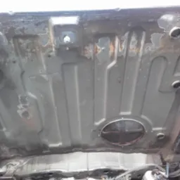 Podwozie samochodu z widocznymi śladami korozji i miejscami po naprawach blacharskich, widoczne otwory technologiczne i elementy konstrukcyjne.