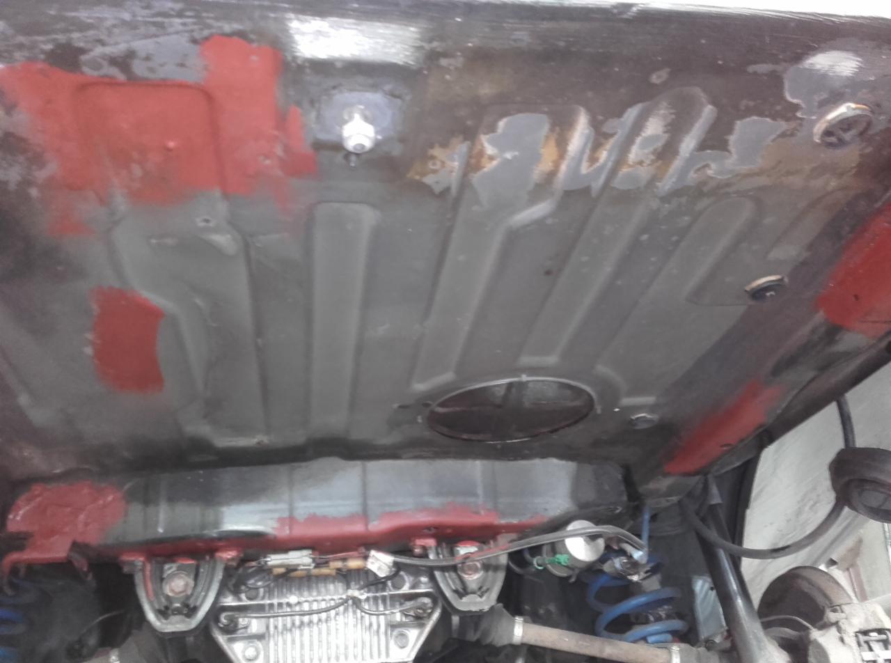 Podwozie samochodu w trakcie renowacji: widoczne ślady korozji, fragmenty nałożonej czerwonej farby podkładowej, elementy zawieszenia i układu wydechowego.