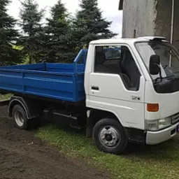 Biała ciężarówka z niebieską skrzynią ładunkową stoi na trawiastym podłożu przed budynkiem i drzewami.