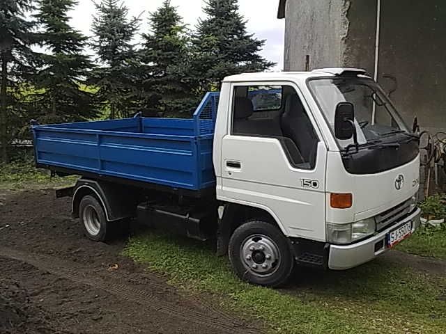 Biała ciężarówka z niebieską skrzynią ładunkową stoi na trawiastym podłożu przed budynkiem i drzewami.