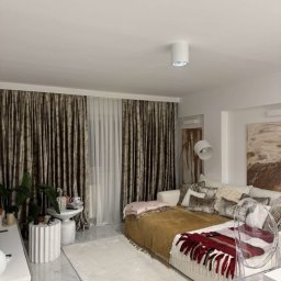 Wid-Art Studio Dekoracji Okien - Elegancki salon z ciężkimi, marszczonymi zasłonami w brązowo-złoty wzór i białymi firanami. Duża beżowa sofa narożna z pledami i poduszkami, biały dywan na błyszczącej, marmurowej podłodze.