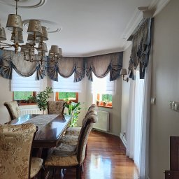 Wid-Art Studio Dekoracji Okien - Elegancka jadalnia z klasycznym stołem i krzesłami. Okna zdobią szaro-niebieskie zasłony wiedeńskie z białymi roletami rzymskimi, a sufit ozdobny żyrandol.