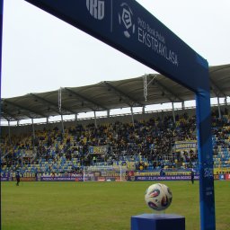Krzysztof Szymański - Piłka nożna na niebieskim postumencie na murawie stadionu, z trybunami pełnymi kibiców i niebieskim banerem PKO Bank Polski Ekstraklasa w górnej części kadru.