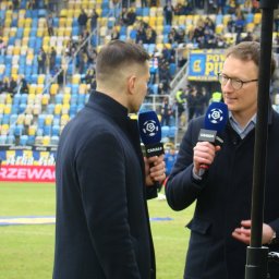 Krzysztof Szymański - Dwaj reporterzy z mikrofonami Canal+ z logo Ekstraklasy prowadzą wywiad na murawie stadionu piłkarskiego, w tle trybuny z kibicami.