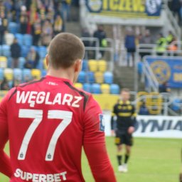 Krzysztof Szymański - Piłkarz w czerwonej koszulce z nazwiskiem WĘGLARZ i numerem 77 oraz napisem SUPERBET na plecach, stojący tyłem na zielonej murawie stadionu, z rozmytymi trybunami w żółto-niebieskich barwach...