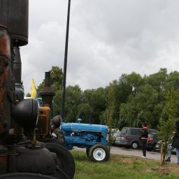 Krzysztof Szymański - Częściowo widoczny, zardzewiały traktor na pierwszym planie, obok błękitny, odrestaurowany Fordson Major na trawie. W tle parking z samochodami i dwie osoby.