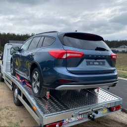 Autolaweta - Granatowy Ford Focus kombi bezpiecznie transportowany na srebrnej platformie w Podolu, zabezpieczony czerwonymi pasami, na tle lasu i domów.