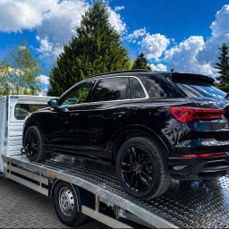 Autolaweta - Czarny SUV Audi Q3 z tablicą CAL 2UT6 transportowany na białym samochodzie pomocy drogowej, zaparkowanym na bruku w słoneczny dzień z błękitnym niebem i drzewami.
