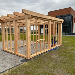 PPHU DREWMAR Zbigniew Jangas - Szkielet drewnianej pergoli lub altany z jasnego drewna, z widocznymi rurami instalacyjnymi, na szarym bruku obok nowoczesnego budynku.