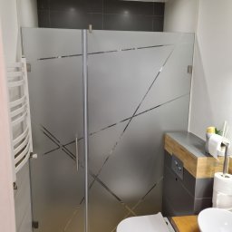 Paweł Dindorf - Nowoczesna łazienka z kabiną prysznicową z matowego szkła z geometrycznym wzorem, obok podwieszana toaleta i biały grzejnik drabinkowy.