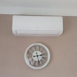GPtermoinstal - Biała jednostka klimatyzatora ściennego AUX na beżowej ścianie, z dekoracyjnym zegarem z rzymskimi cyframi poniżej. Widoczna lampa industrialna i łapacz snów.