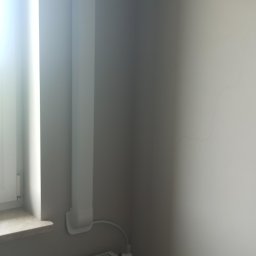 GPtermoinstal - Biała jednostka klimatyzacji ściennej zamontowana w rogu pomieszczenia, z estetycznym korytem instalacyjnym biegnącym wzdłuż ściany obok okna i grzejnika.