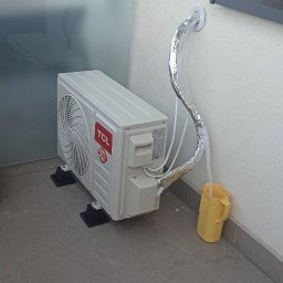 GPtermoinstal - Zewnętrzna jednostka klimatyzacji TCL na szarych płytkach balkonu, podłączona do ściany izolowanymi rurami, z odpływem skroplin do żółtego wiadra. Odbicie w szklanej balustradzie.