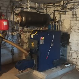 GPtermoinstal - Kotłownia z niebieskim piecem na pellet Bioline i podajnikiem paliwa, z czerwonym naczyniem wzbiorczym i czarnym zbiornikiem buforowym na tle białych ceglanych ścian.
