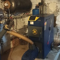 GPtermoinstal - Niebiesko-żółty kocioł grzewczy ZEBIEĆ 10 kW z podajnikiem, podłączony do instalacji z czarnym zbiornikiem wyrównawczym i niebieskim naczyniem przeponowym, w kotłowni.