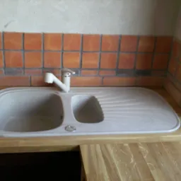 Dwukomorowy zlewozmywak z beżowego kompozytu, zamontowany w drewnianym blacie, z baterią w kolorze ecru i obudowany ścianą z pomarańczowych płytek kwadratowych z ciemną fugą.