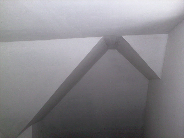 Szare wnętrze z widoczną konstrukcją sufitu podwieszanego, tworzącą geometryczny wzór z ukośnych linii i kątów prostych.