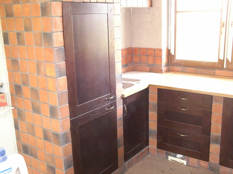 Wykończona kuchnia z ciemnymi drewnianymi szafkami i pomarańczowo-brązowymi płytkami na ścianach, z widocznym fragmentem okna i blatem roboczym, w trakcie lub po remoncie.