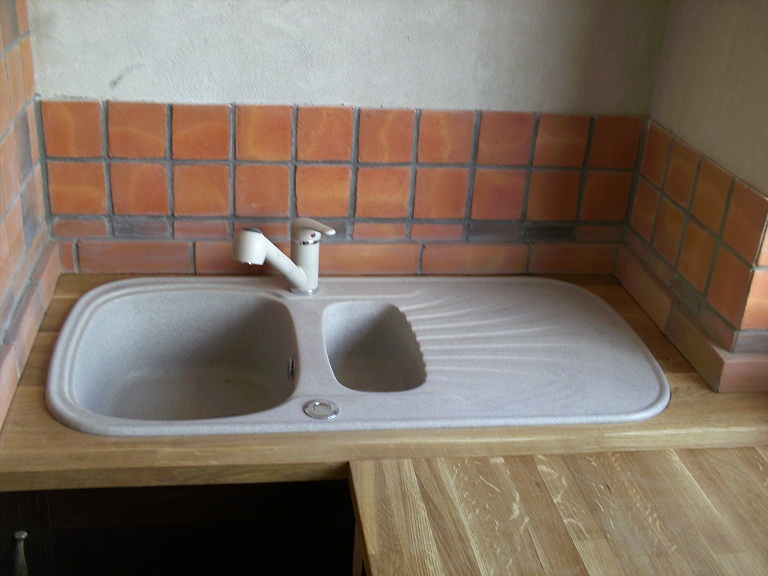 Dwukomorowy zlewozmywak z beżowego kompozytu, zamontowany w drewnianym blacie, z baterią w kolorze ecru i obudowany ścianą z pomarańczowych płytek kwadratowych z ciemną fugą.