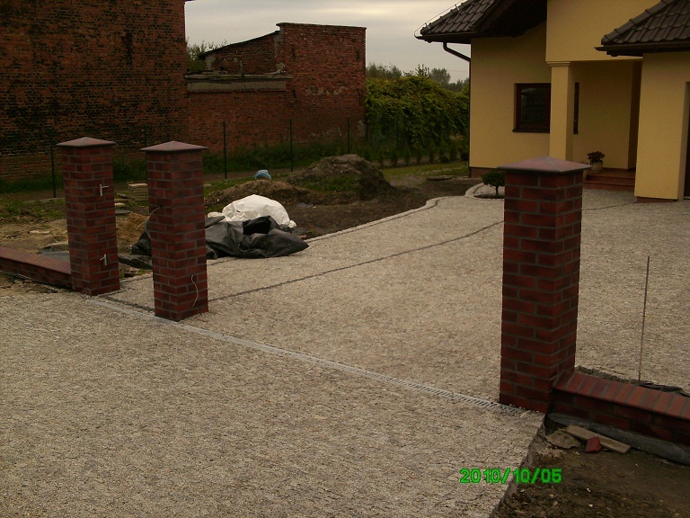 Klinkierowe słupki ogrodzeniowe z przyłączami elektrycznymi na żwirowym podłożu, w tle dom jednorodzinny i budynek gospodarczy z czerwonej cegły.