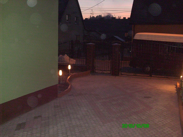 Klinkierowe ogrodzenie z przesuwną bramą, oświetlone niskimi lampkami, prowadzące do posesji z budynkiem w tle, sfotografowane o zmierzchu.