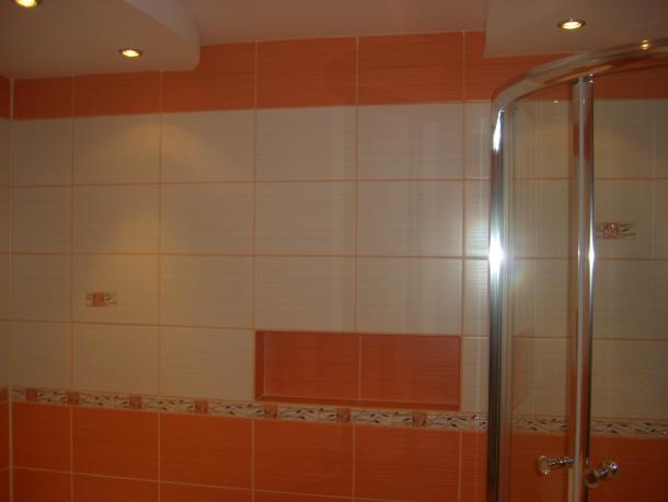 Wykończona łazienka z pomarańczowo-białymi płytkami, widoczna kabina prysznicowa z chromowanymi uchwytami i wnęka w ścianie wyłożona pomarańczowymi płytkami.