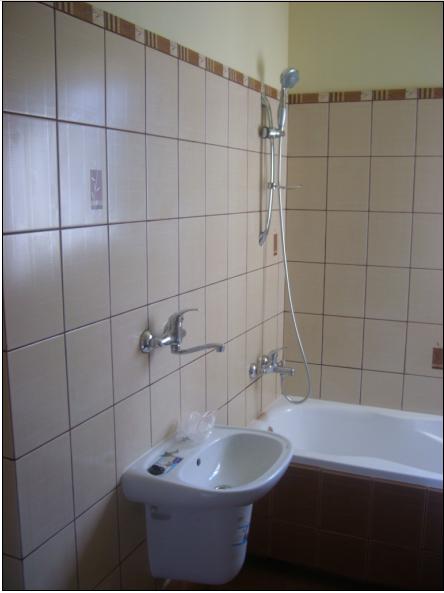 Wykończona łazienka z beżowymi płytkami, widoczna umywalka z baterią, wanna z prysznicem oraz dekoracyjny pasek płytek pod sufitem.