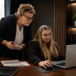 Scutum Group Polska - Dwie kobiety w okularach analizują dokumenty na srebrnym laptopie HP w eleganckim biurze. Starsza kobieta z markerem nadzoruje pracę młodszej, obok regał z książkami i waga sprawiedliwości.