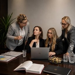 Scutum Group Polska - Cztery kobiety w biznesowych strojach, w tym jedna wskazująca długopisem, intensywnie dyskutują przy laptopie Dell na ciemnym stole. Widoczne są segregatory, otwarta księga i szklany dekanter.