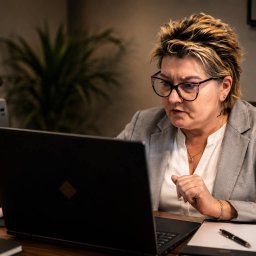 Scutum Group Polska - Skupiona kobieta w okularach i szarym żakiecie intensywnie analizuje ekran laptopa, z dłonią na myszce i zaciśniętą pięścią, w biurze z drukarką HP.