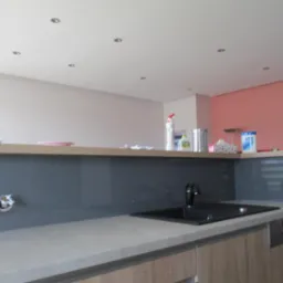 Szara szklana ściana nad blatem kuchennym z czarnym zlewem i baterią, widoczne puszki elektryczne w ścianie, w tle choinka i akcesoria kuchenne.